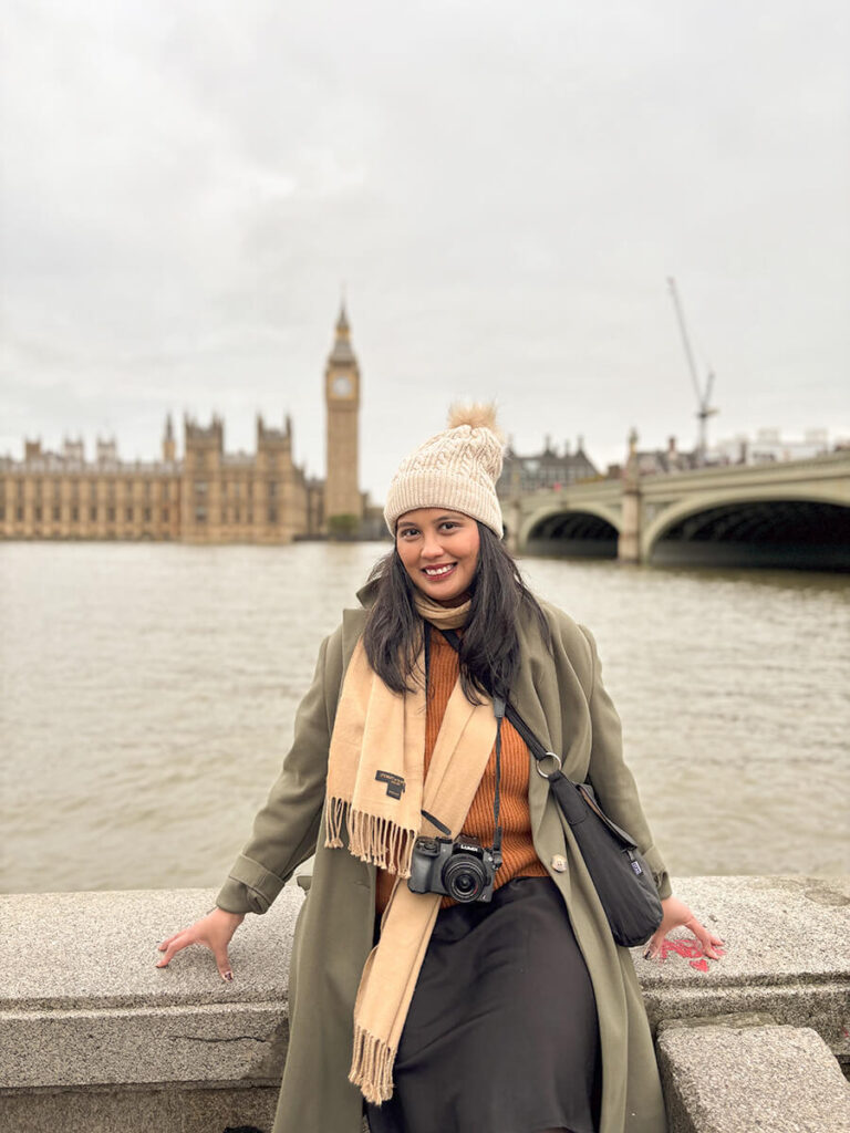Hannah wearing an olive green coat, brown sweater, tan scarf, beige beanie, in front of Big Ben and Westminster Bridge in London