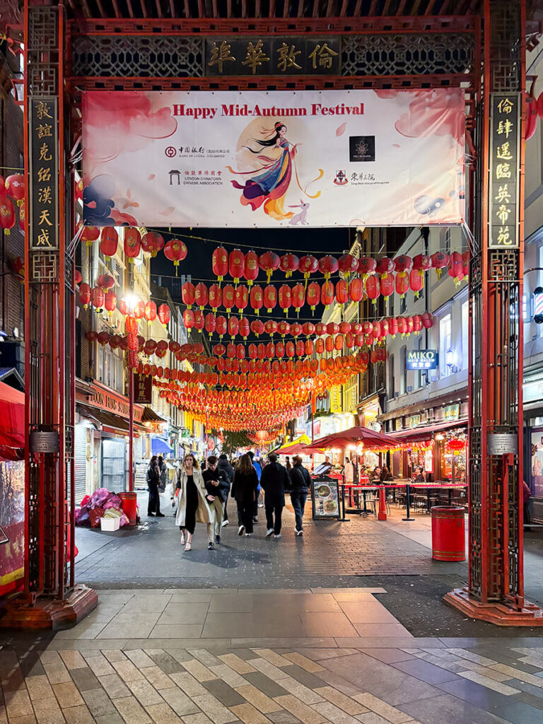 The paifang in Chinatown in London, located in Soho. Right behind the gate, there are rows of red lanterns. There is a large sign that says "Happy Mid-Autumn Festival"