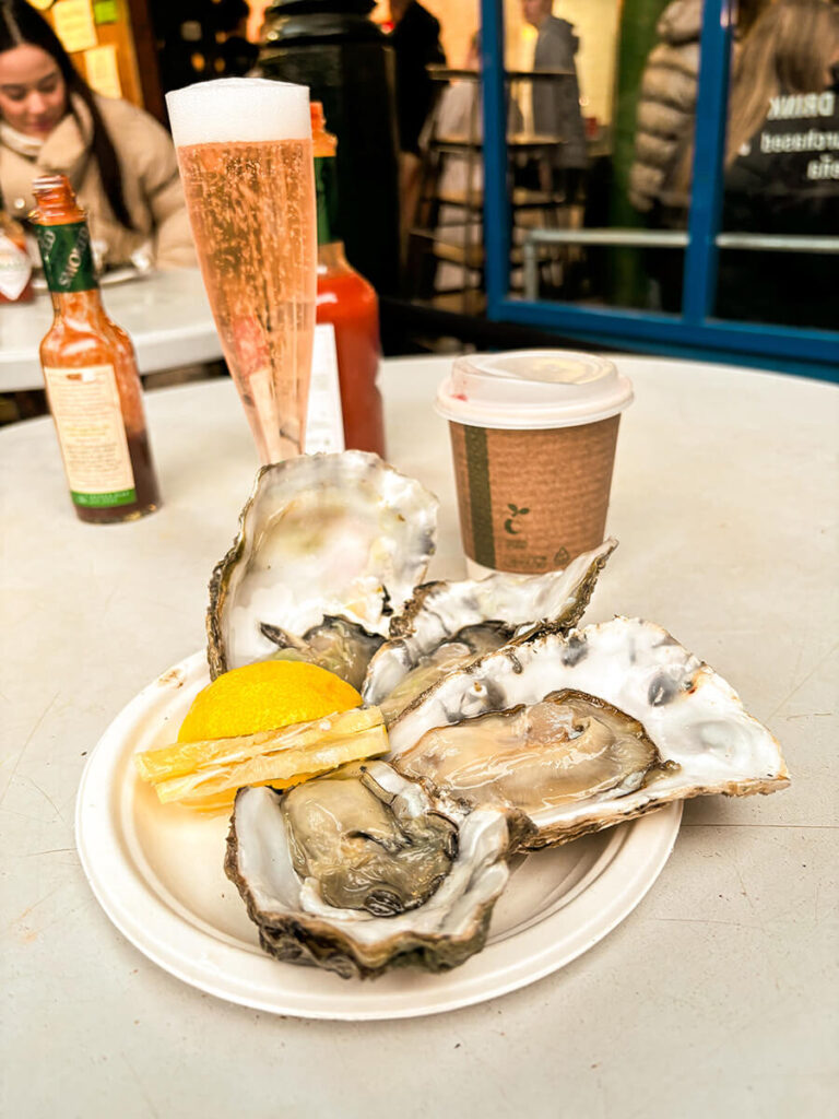 a plate of four oysters with a slice of lemon. Behind the plate is a cup of mulled wine and a glass of rose, with some hot sauces. These oysters were from Richard Haward Oysters, a stall in Borough Market in London