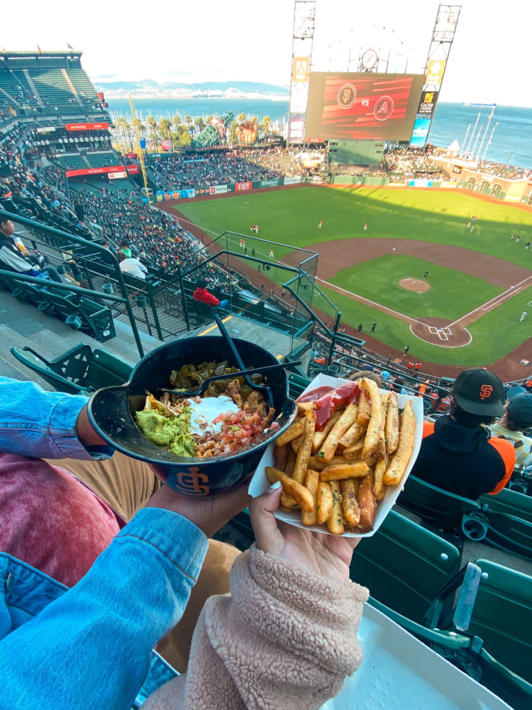 Hannah and Cecilio holding a tray of fries and a plastic hat of nachos during a San Francisco Giants Game at Oracle Park