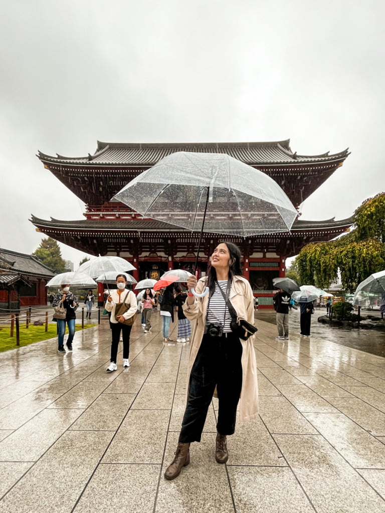 Woman wearing a beige trench coat, black and white striped shirt, black crossover bag, black pants, and brown boats, holding an clear umbrella on a rainy day. Behind her is the Sensoji Temple, a Buddhist temple in Tokyo. There are other people walking behind her holding umbrellas.