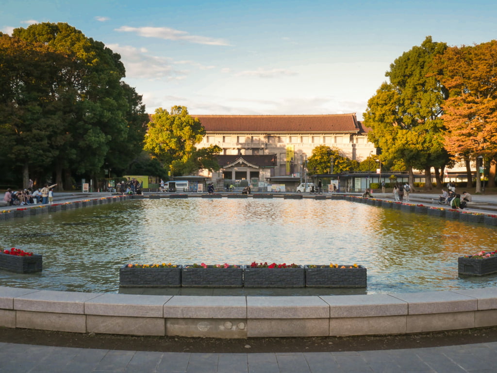 large pond in front of the Tokyo National Museum located in Ueno Park, taken on a sunny day during fall with trees on both sides. The sky is sunny with clouds just before the sun is setting