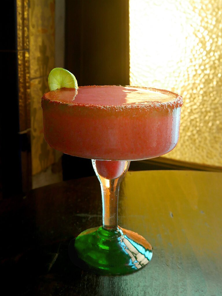 A large margarita glass filled with a pink strawberry margarita and salt and tajin rim. On the left there is a small lime on the rim. The margarita is on a table, with a window behind it. This is in Bar Andaluz in Ensenada, Mexico, the birthplace of the margarita drink