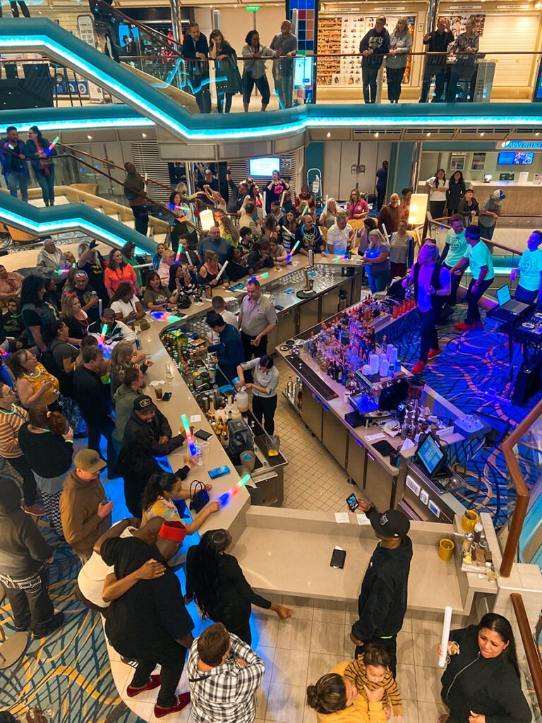 Passengers on the atrium in Carnival Radiance cruise filling the atrium and staircase to celebrate 80's Rock and Glow night, one of the themed parties at night in Carnival cruise ships. Some passengers are wearing regular clothes, and some are wearing 80's-inspired outfits with neon tops, crimped hair, and scrunchies. Some passengers are holding a rainbow glow-in-the-dark wand. They are all dancing to live music featuring 80's hits, and the cruise director is in front of the band encouraging the passengers to party. In between the passengers and the band is the bar, with bartenders making drinks for passengers