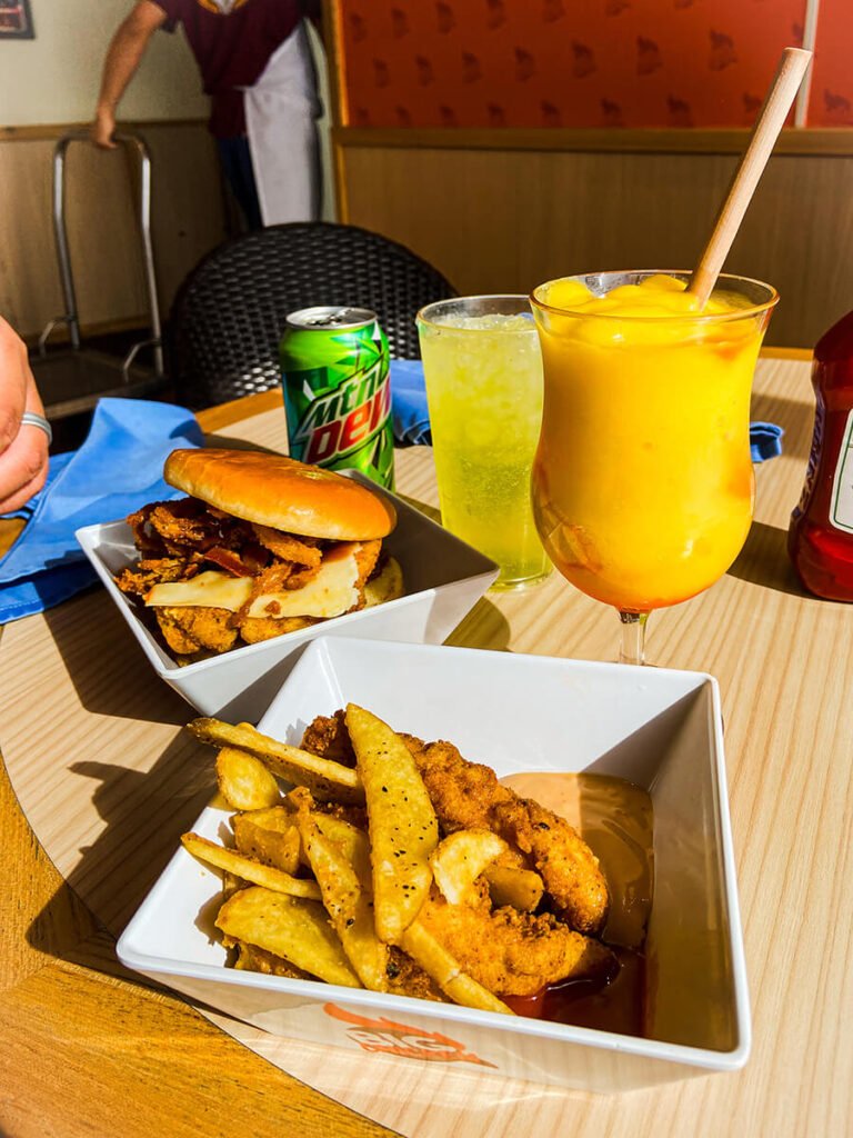 Two bowls full of food from Big Chicken, Shaquille O'Neal's eatery in Carnival cruise ships. On the front, there is fried chicken strips and french fries. Behind that is The Big Aristotle, a sandwich with muenster cheese, fried onions, bacon, and BBQ sauce. The drinks shown are an empty can of Mountain Dew with a cup of the drink poured from the can, and a cocktail called Kiss on the Lips, which has peach schnapps, mango puree, and grenadine.