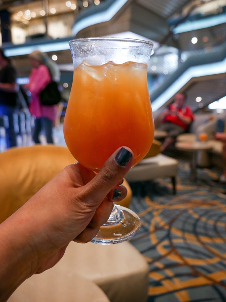 Hannah's hand holding a Pirate Punch, a cocktail on Carnival Radiance cruise in the atrium. The cocktail is a bright orange/peach flavor, and has 2 types of rum, cranberry, amaretto, pineapple and orange juice. There are two people standing behind the drink and one person sitting down on the lounge. At a distance, there is a staircase leading to the deck above the atrium.