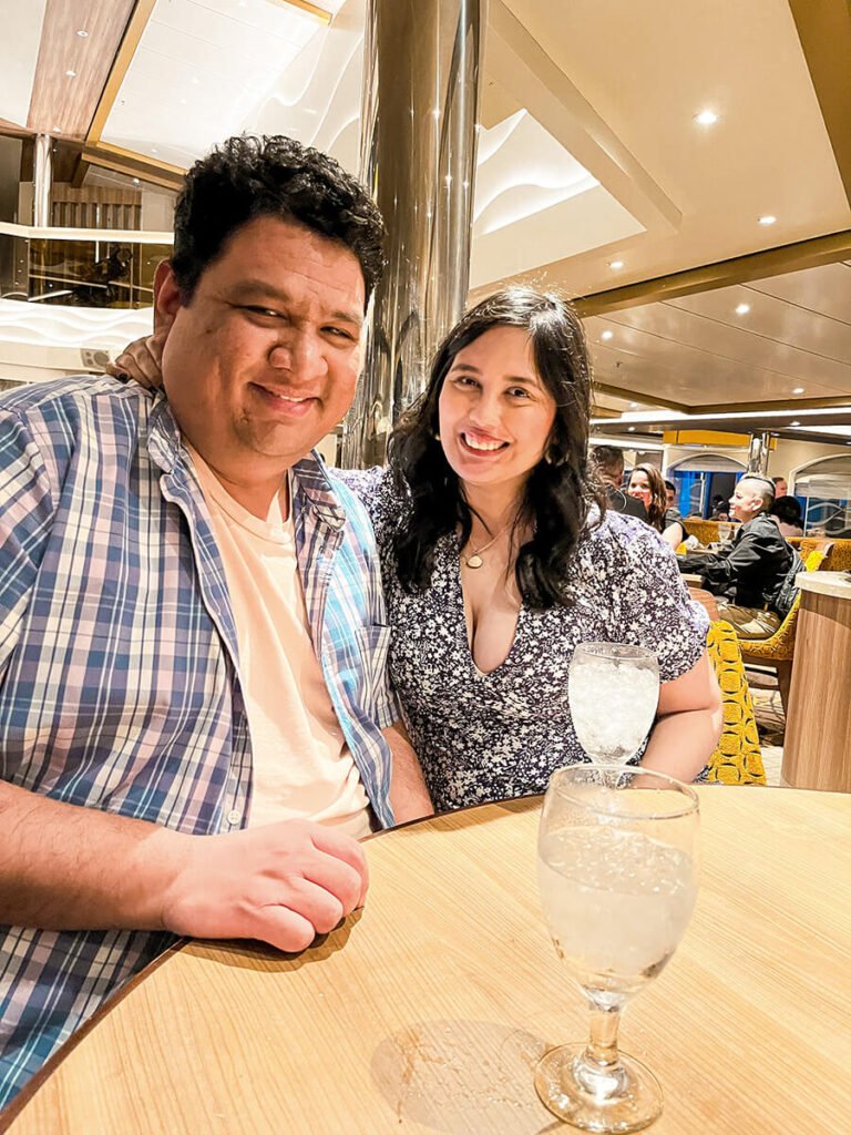 Hannah and Cecilio sitting down posing in front of the camera at the main dining room in Carnival Radiance cruise, with two glasses of water with ice. There are two people behind them sitting down