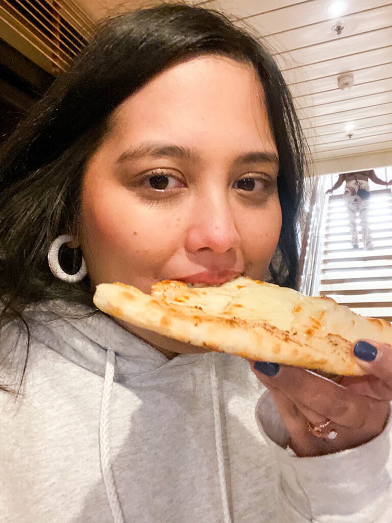 Hannah eating cheese pizza from Pizzeria del Capitano, a pizza eatery from Carnival Radiance and other Carnival cruise ships. There is someone on the staircase behind her