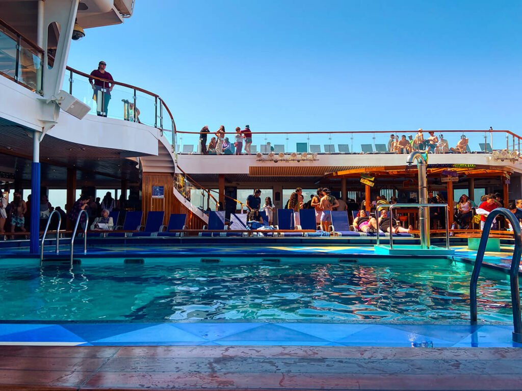 Empty swimming pool on Carnival Radiance cruise on the lido deck on a bright and sunny day. At a distance, there are some people sitting on the beach chairs, and other people lining up to get lunch. There are some people hanging out on the beach chairs on the deck above the lido deck.