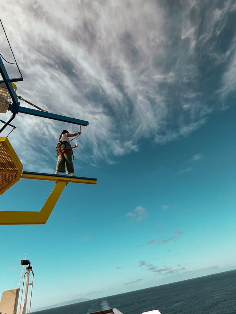 Hannah walking on a yellow plank, wearing a vest strapped to a blue plank above, overlooking the ocean. She is doing the ropes course in a Carnival Radiance ship, a fun activity where you walk on different obstacles to test your balance. For safety reasons, you are strapped the whole time.