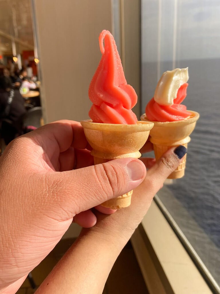 Two people's hands holding an ice cream cone in Carnival Radiance cruise ship. On the left, the cone has pink strawberry frozen yogurt. On the right, the cone has strawberry frozen yogurt with vanilla soft serve ice cream. On the right in the background is a window overlooking the sea, and the left there are people sitting down eating at the food areas.