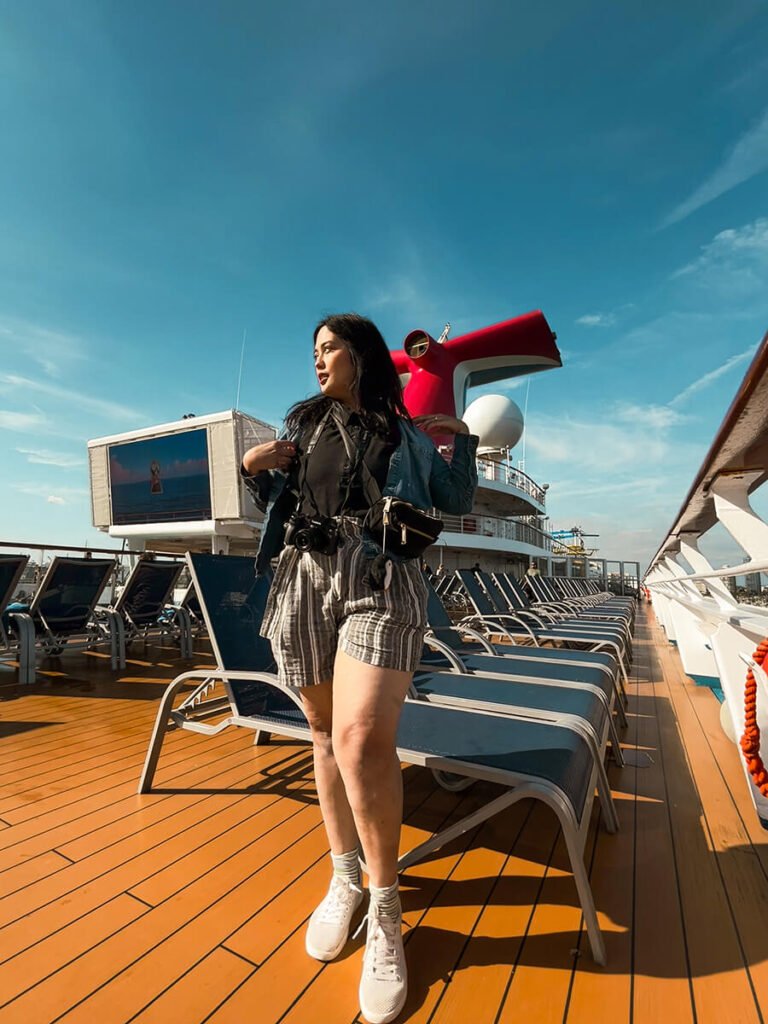 Hannah standing above the lido deck on the Carnival Radiance cruise on a sunny day, right before the sailaway party. There are two rows of empty beach chairs right behind her. At a distance, there is Carnival's signature red wing which directs the exhaust