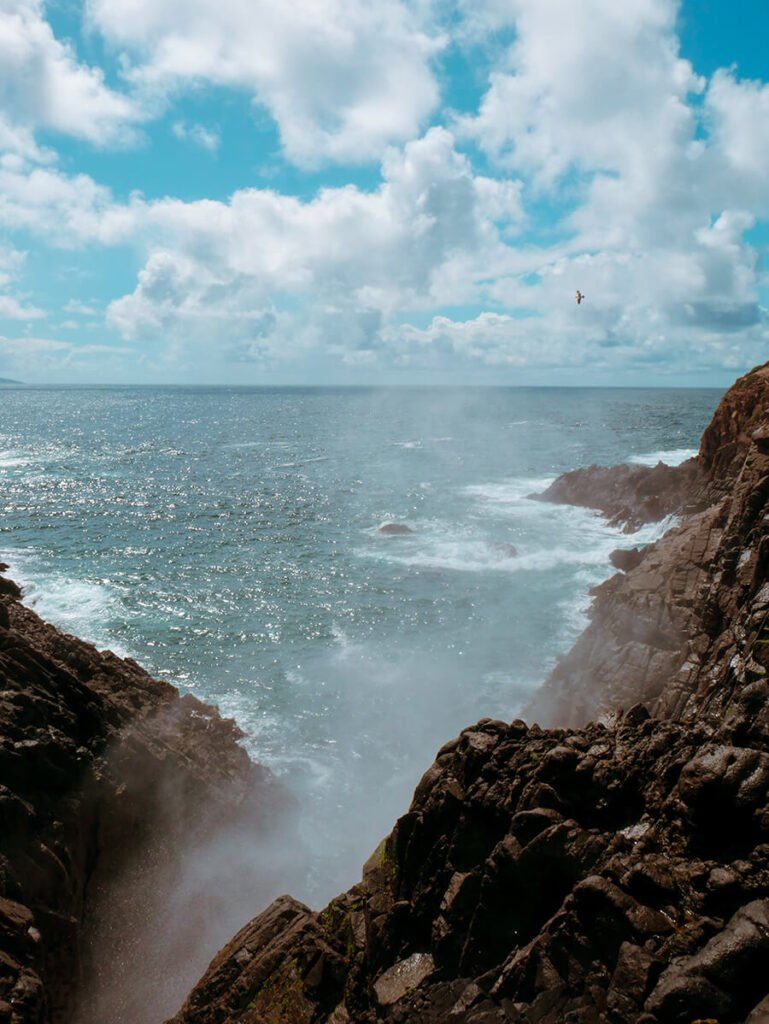 a large mist of water spouting from the Pacific Ocean at a distance on a sunny day with clouds. This is La Bufadora, one of the largest blowholes in the world, located on the Punta Banda Peninsula, 45-60 minutes south of Ensenada, Mexico. The rock formations next to it are the seacaves that allow the water to erupt.