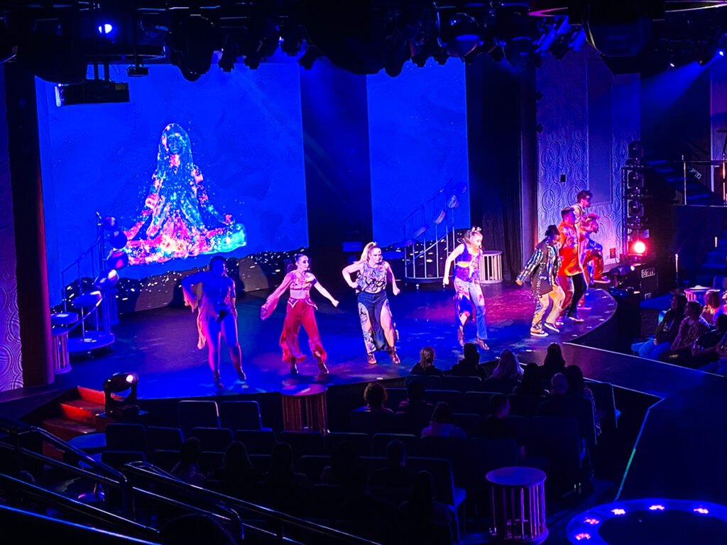 Seven performers singing and dancing on stage at the Liquid Lounge on the Carnival Radiance ship. They are performing to Rock Revolution, a musical show featuring 60s hits. The lighting all around the area is blue, red, and purple, with the passengers watching being completely dark.