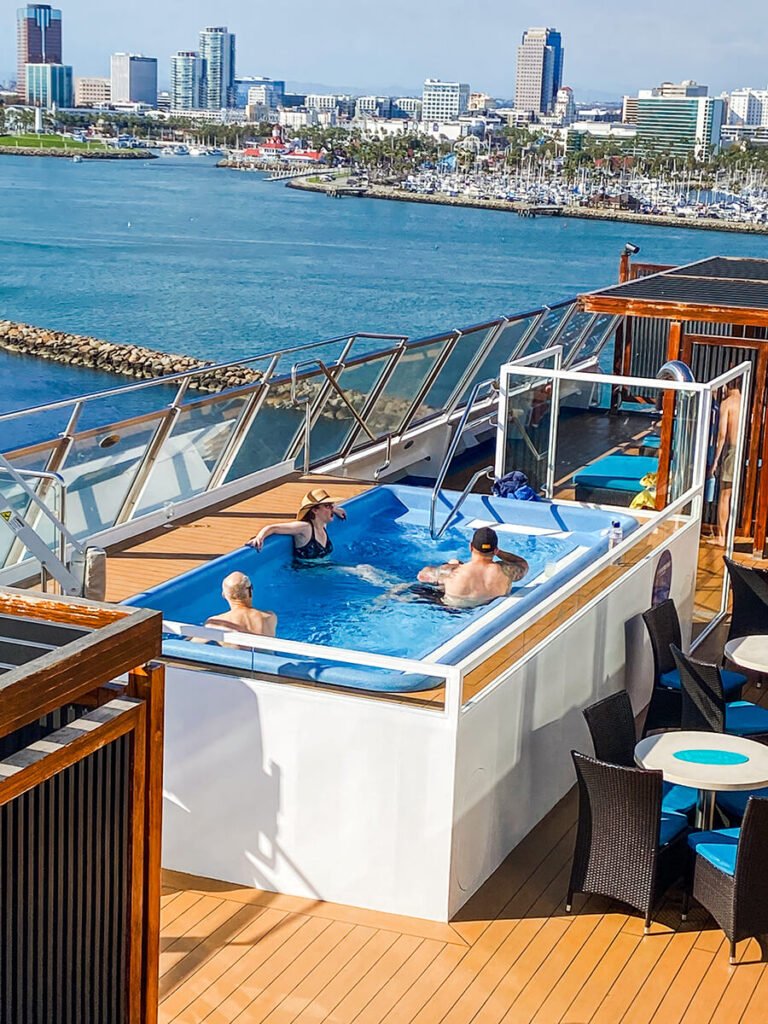 Jacuzzi in Serenity Deck, the adults-only area on the 14th deck on the Carnival Radiance cruise ship on a sunny day. There are 3 people in the jacuzzi, with some empty chairs. The deck is overlooking the port of Long Beach, where Carnival Radiance sails out of