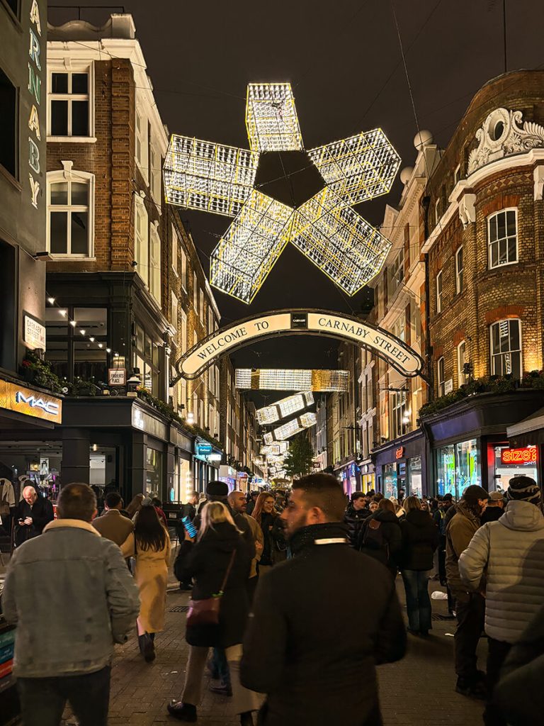 View of Carnaby Street in London at night with Christmas lights and shapes hanging, and a crowd of people in the middle of the street