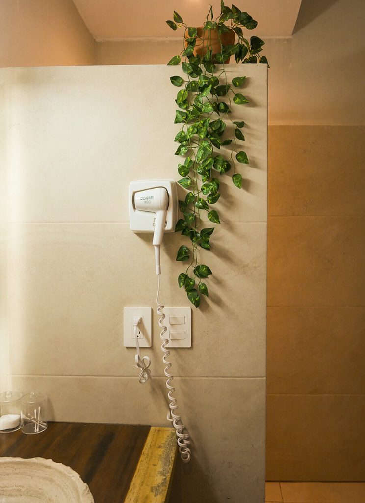 Bathroom of Casa Victoria Malecon, an adults-only guesthouse in Puerto Vallarta. Pictured is a white hairdryer and some sockets. there is also a plant with vines and leaves hanging loose. Behind the wall, there is a toilet (not shown)