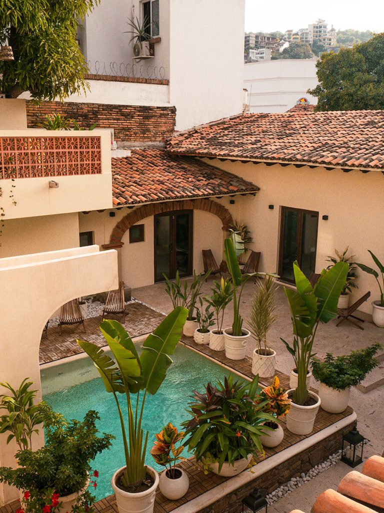 Exterior of Casa Victoria Malecon, an adults-only guesthouse in Puerto Vallarta. Outside, there is a pool surrounded by plants. There are brick roofs. The view is taken from the second floor. You can see views of buildings atop hills at a distance