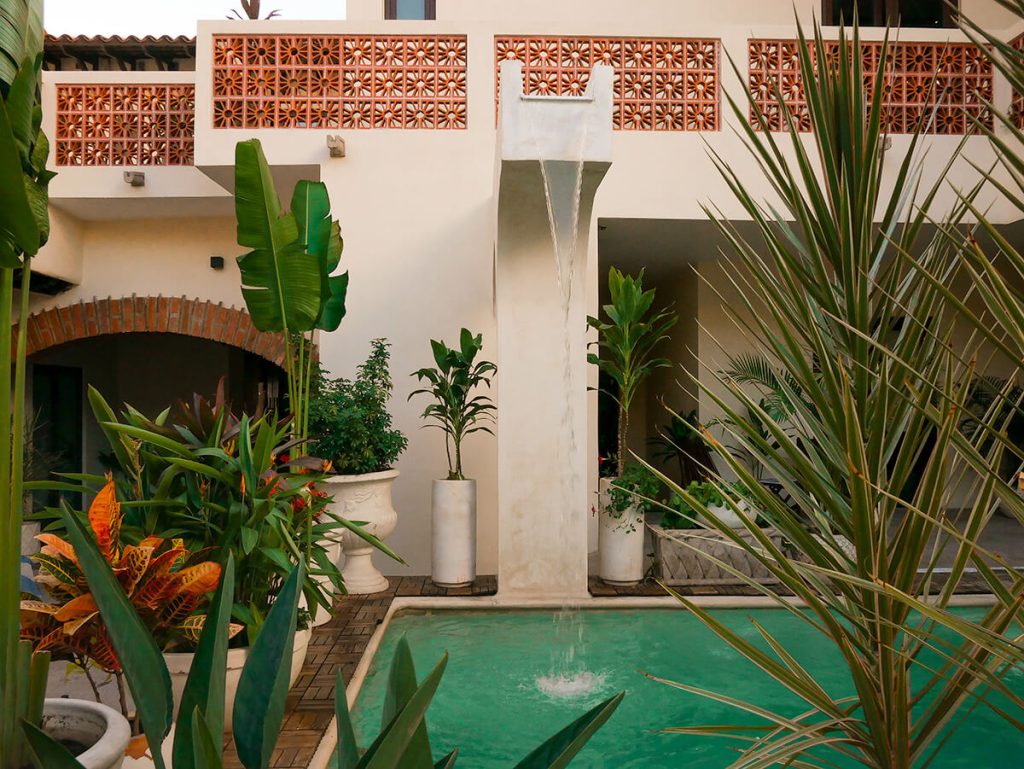 Pool area in Casa Victoria Malecon, an adults-only guesthouse in Puerto Vallarta, Mexico. The pool is surrounded by plants, and there is a small fake waterfall above it that also can turn off. The building is a cream white with some terracotta bricks.