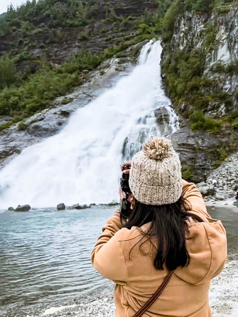 Hannah wearing a beige knitted beanie and a tan jacket taking a photo from behind of the Nugget Falls located in Mendenhall Glacier in Juneau, Alaska