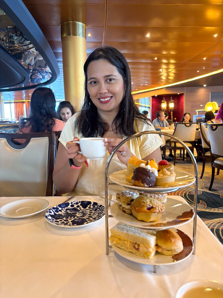Hannah holding a cup of tea during afternoon Dutch tea on Holland America Zaandam. In front of her are a tier of Dutch sandwiches, pastries, and desserts.