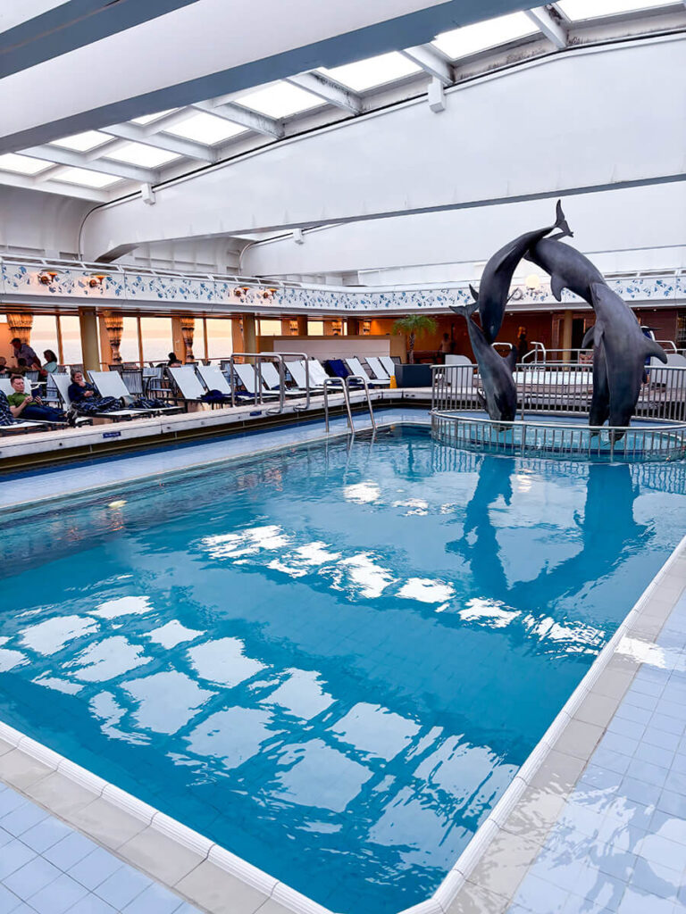 Lido pool on Holland America Zandam. Behind the pool, there's an arch structure of dolphins, and two jacuzzis. The pool has a retractable roof.