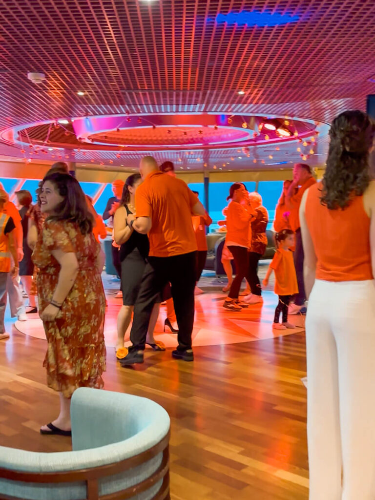 Passengers on Holland America Zaandam on the dance floor in Crow's Nest Bar, decked out in orange outfits for the Orange Party. The lighting is orange too