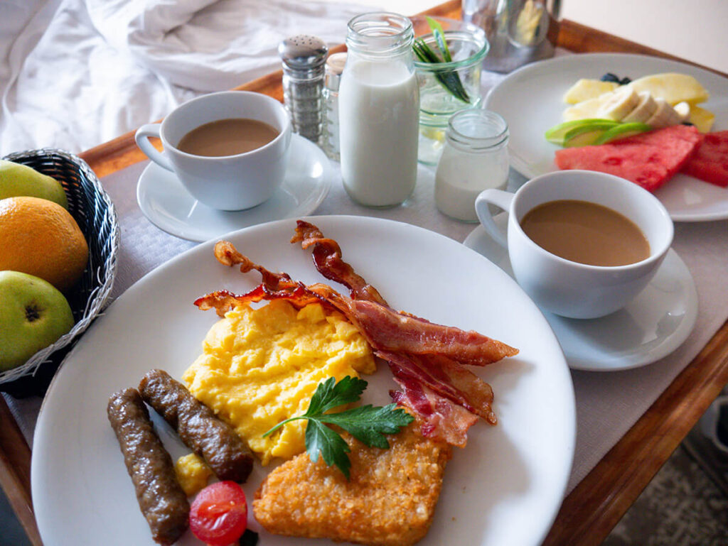 breakfast room service on Holland America Zaandam. On the front, there's a plate with 2 sausages, scrambled eggs, hash browns, a tomato, and bacon. To the left, there is an orange and 2 apples. Behind the plate of eggs and meats are two coffees, a glass of milk and cream. To the right, there is a bowl of fruit featuring watermelon, bananas and blueberries.