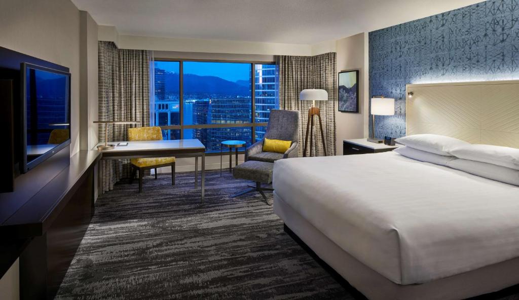 A King-sized bed in Hyatt Regency Vancouver, with a ceiling-to-floor window overlooking Downtown Vancouver at night. There is also a desk and some lamps