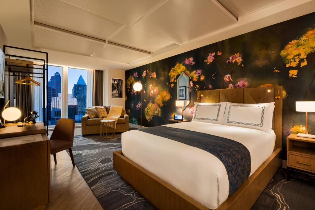 Queen sized bed in EXchange hotel window in a suite. Behind the bed is floral wallpaper. There are ceiling-to-floor windows overlooking Downtown Vancouver. There is a closet, couch, side table on each side of the bed, and a desk