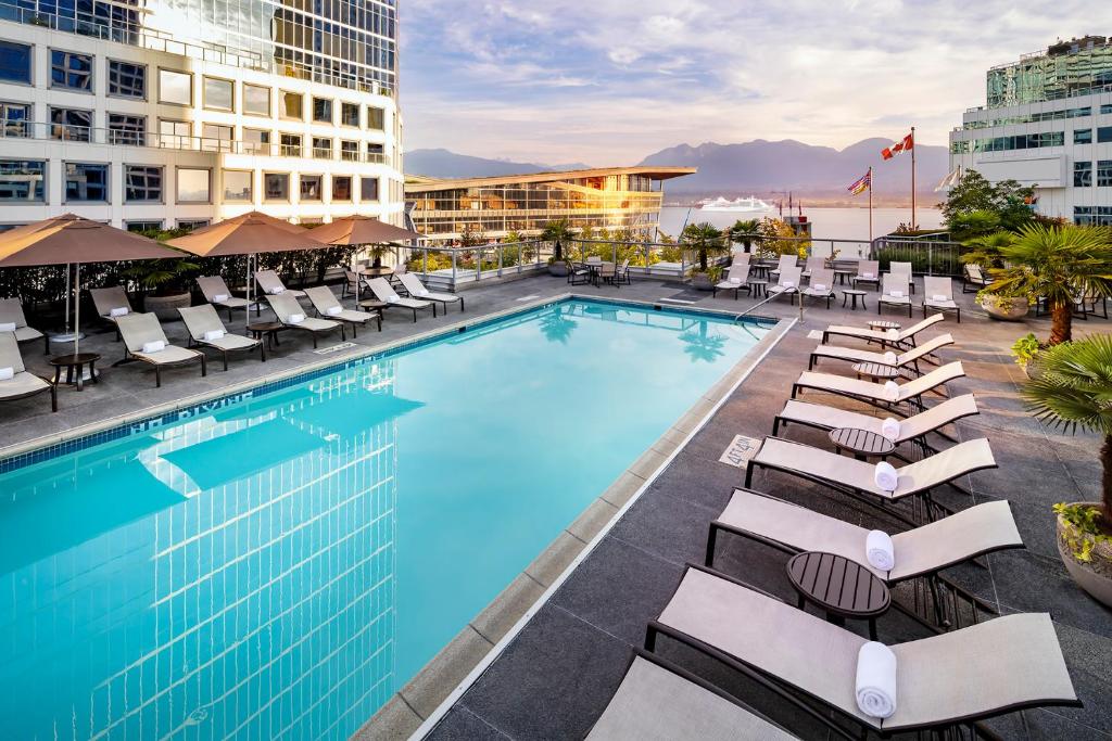 Rooftop pool in Fairmont Waterfront in Vancouver, overlooking Canada Place and the Harbor. There are rows of beach chairs on each side of the pool, and trees lined