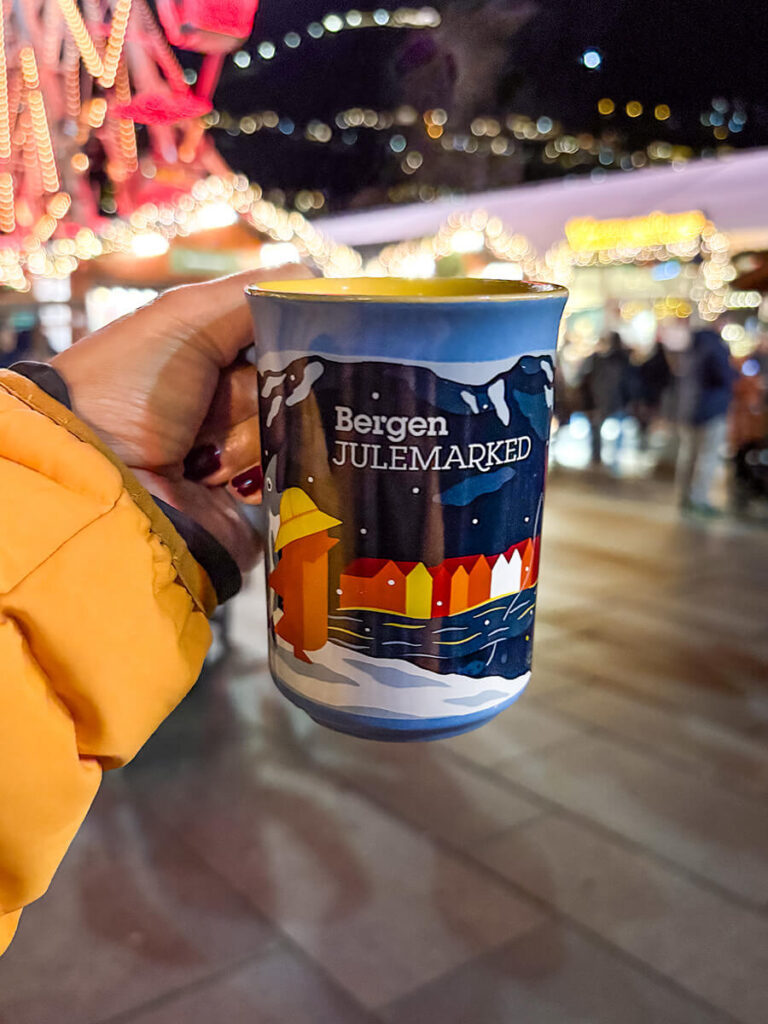 Hannah holding a mug at the Christmas market in Bergen, Norway. The mug is blue with an illustration of Bryggen, Mount Floyen, and the fjord. Text in the mug says "Bergen Julemarked"