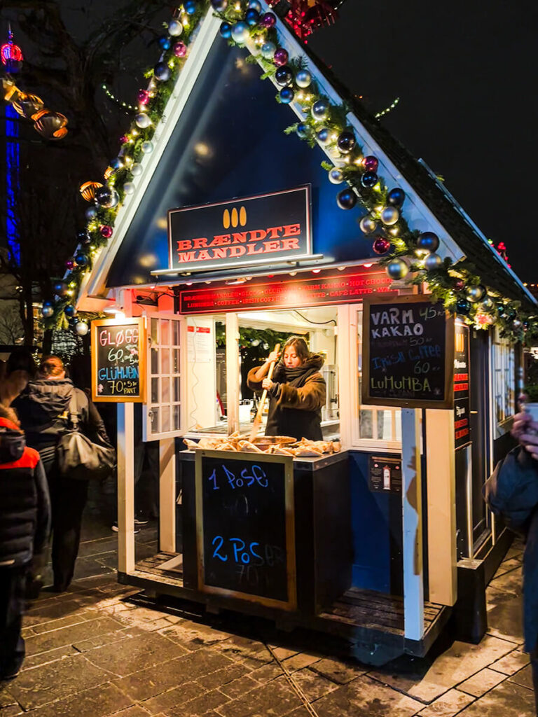 Woman at Tivoli Gardens in Copenhagen at night during Christmas season, in a stall that says "Brændte Mandler", roasting almonds. The stall roof is lined with garlands and ornaments. The stall also sells glogg and hot cocoa.