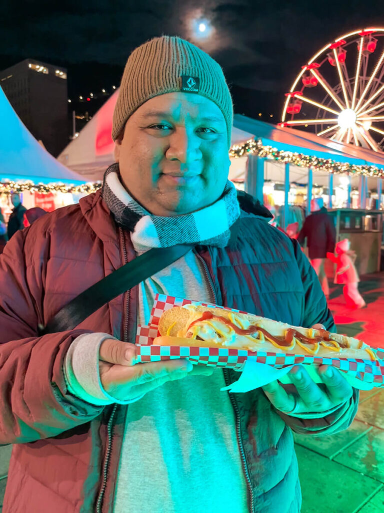 Cecilio holding a hot dog at Bergen Julemarked, the Christmas Market in Bergen at night. Behind him are some stalls and the Ferris wheel