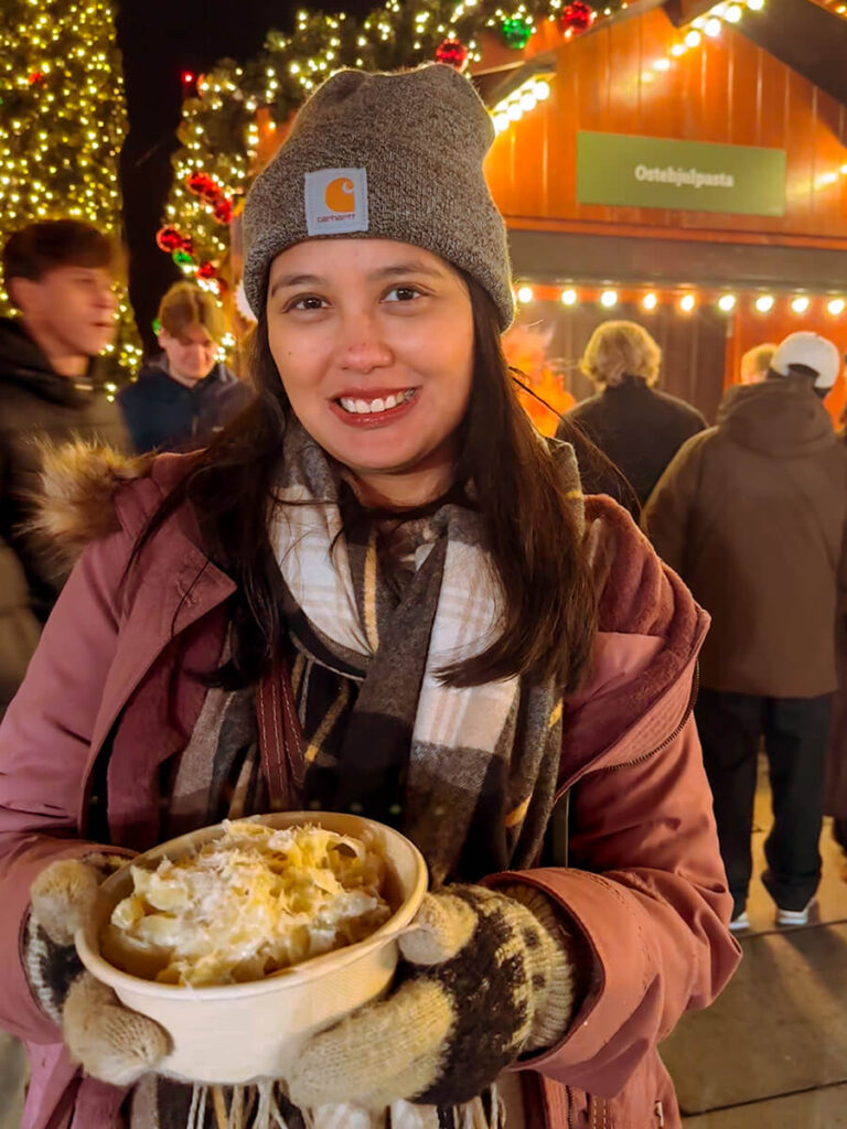 Hannah holding a bowl of pasta at the Bergen Christmas market. Behind her are a few people in line getting the same thing at the stall that says ostehjulpasta, which is where she got the pasta from. ostehjulpasta means cheese wheel pasta, so the pasta was cooked with a cheese wheel