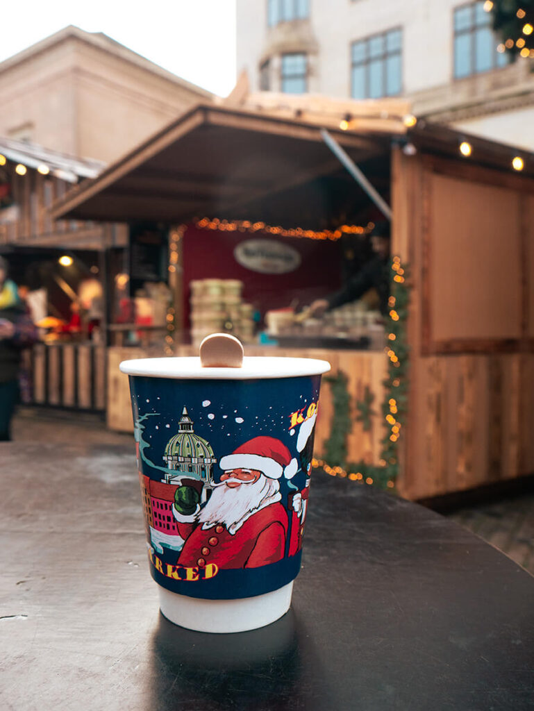 A cup of glogg at the Nytorv Christmas market in Copenhagen. The cup is dark blue with an image of santa, and right behind the cup is a wooden stall