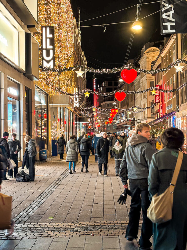 the street Købmagergade in Copenhagen decked out in Christmas lights, hearts, and garlands. There is a crowd of people walking the street at night. On the left is the department store, ILLUM, covered in hanging lights