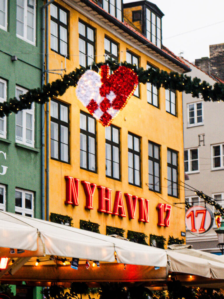 A closeup shot of Nyhavn in Copenhagen during the day in December. There is a Christmas hanging garland with the red and white woven heart in the center called the julehjert, with the yellow Nyhavn 17 building behind