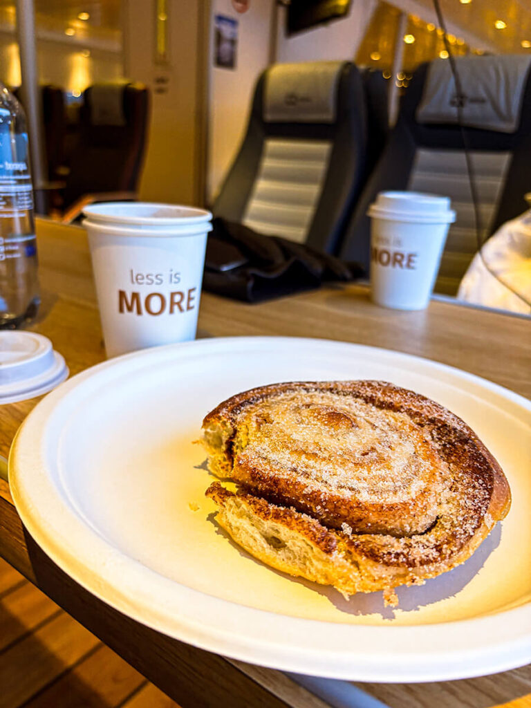 Skillingsboller, a Norwegian cinnamon roll on a paper plate. There are 2 paper cups that says "less is more", and some seats. This was in the Rødne Rygersol boat for the Mostraumen fjord cruise